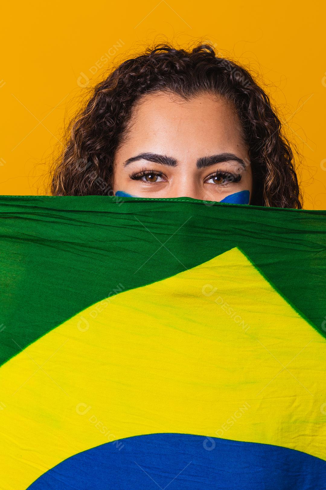 Fã negra misteriosa segurando uma bandeira do Brasil na sua cara. Cores do Brasil ao fundo, verde, azul e amarelo