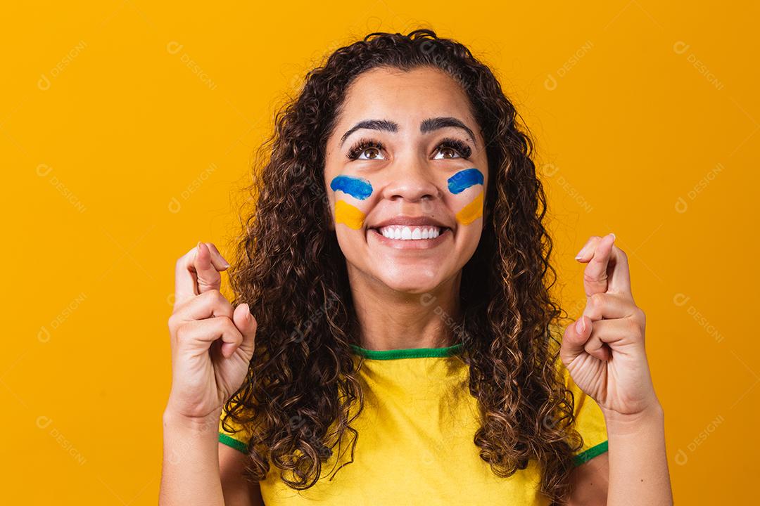 fã brasileiro com dedos cruzados vibrando sorte em fundo amarelo
