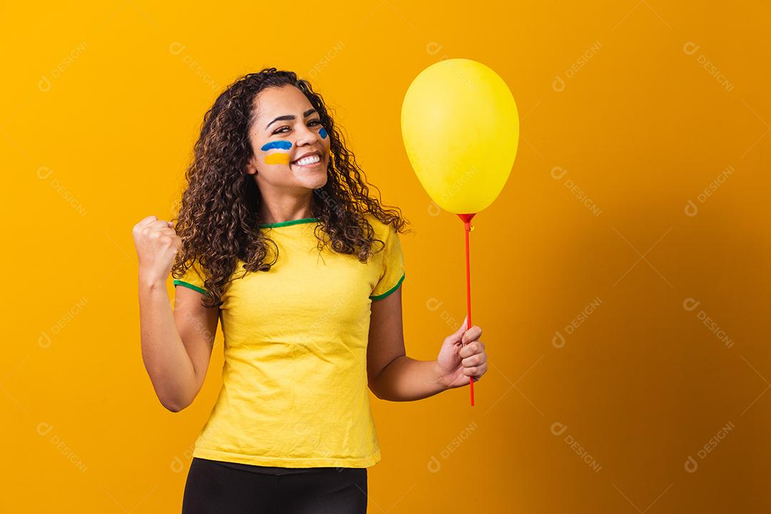 Torcedora brasileira segurando um balão em fundo amarelo