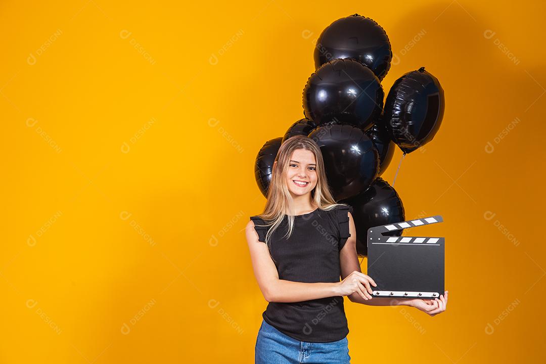 Jovem mulher rindo segurando filme preto clássico fazendo claquete e comemorando com balões de ar preto isolados