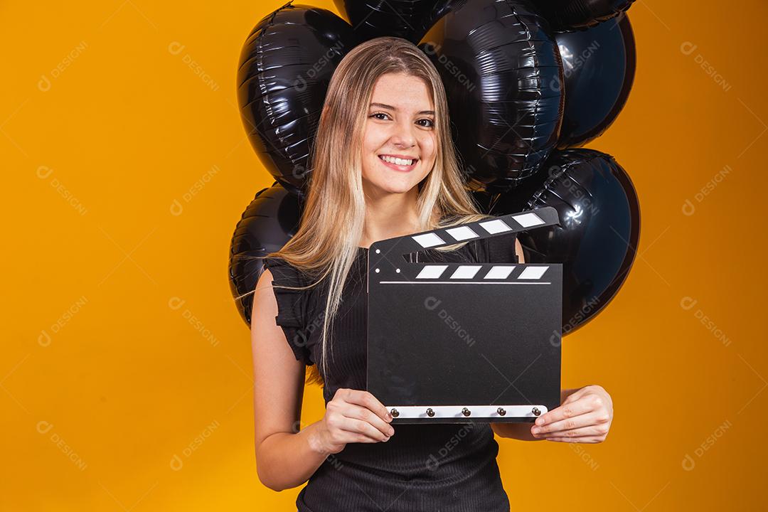 Jovem mulher rindo segurando filme preto clássico fazendo claquete e comemorando com balões de ar preto isolados