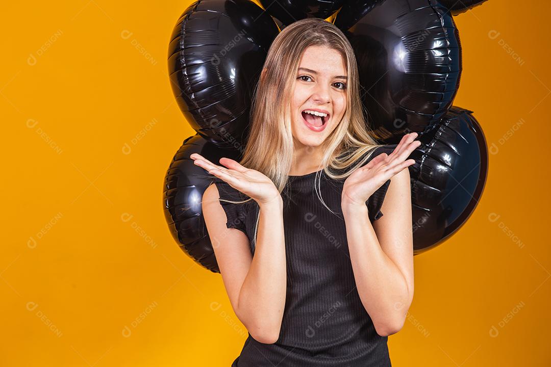 Conceito de sexta-feira negra com mulher atraente loira com linda animada, vestida de preto olha para a câmera fica com fundo amarelo de balões de ar preto, espaço de cópia.