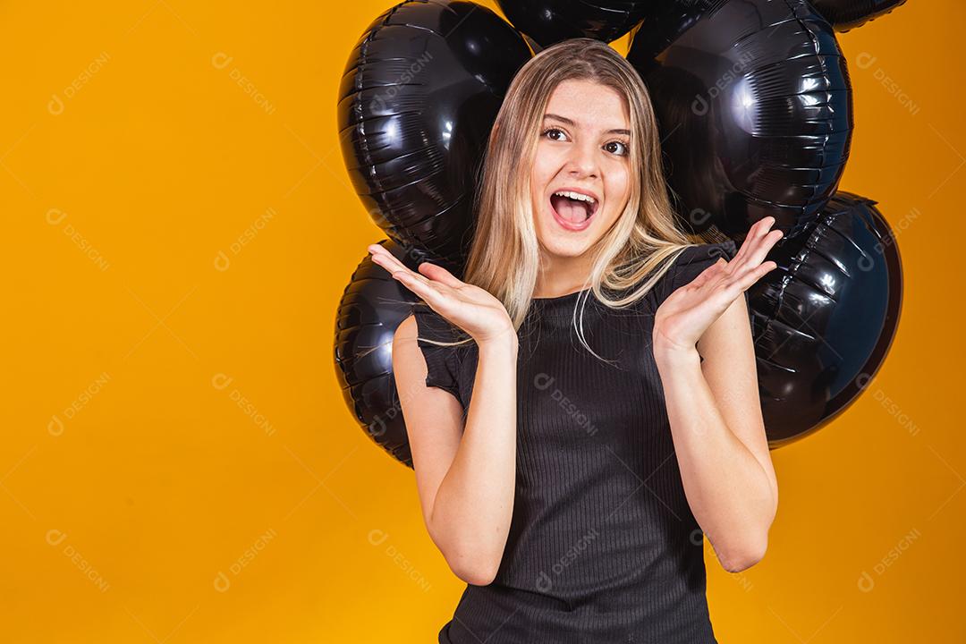 Conceito de sexta-feira negra com mulher atraente loira com linda animada, vestida de preto olha para a câmera fica com fundo amarelo de balões de ar preto, espaço de cópia.