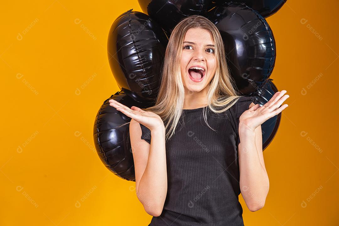 Conceito de sexta-feira negra com mulher atraente loira com linda animada, vestida de preto olha para a câmera fica com fundo amarelo de balões de ar preto, espaço de cópia.
