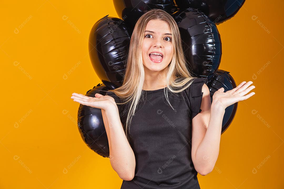 Conceito de sexta-feira negra com mulher atraente loira com linda animada, vestida de preto olha para a câmera fica com fundo amarelo de balões de ar preto, espaço de cópia.