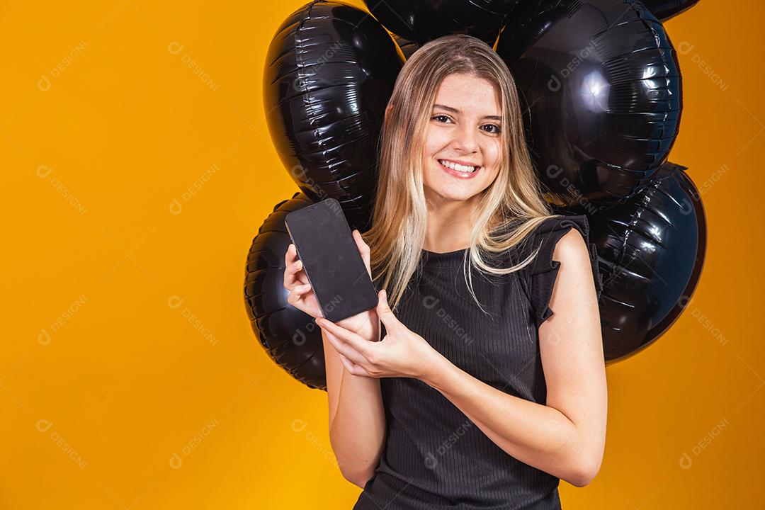 Sorrindo encantadora jovem com celular com tela em branco vazia sobre fundo amarelo com retrato de estúdio de balões de ar. venda sexta-feira negra