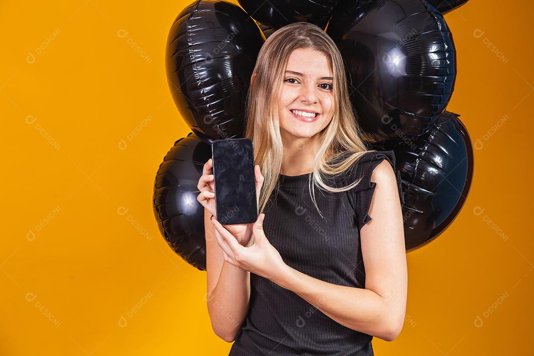 Sorrindo encantadora jovem com celular com tela em branco vazia sobre fundo amarelo com retrato de estúdio de balões de ar. venda sexta-feira negra