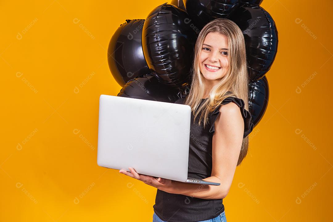 Sorrindo alegre e engraçada morena jovem 20 anos usando computador portátil pc em fundo de cor amarela brilhante com retrato de estúdio de balões de ar. Venda de sexta-feira negra