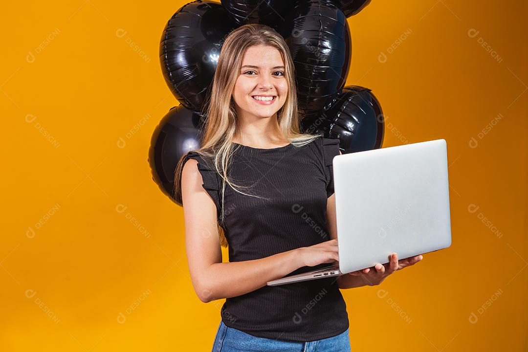 Sorrindo alegre e engraçada morena jovem 20 anos usando computador portátil pc em fundo de cor amarela brilhante com retrato de estúdio de balões de ar. Venda de sexta-feira negra