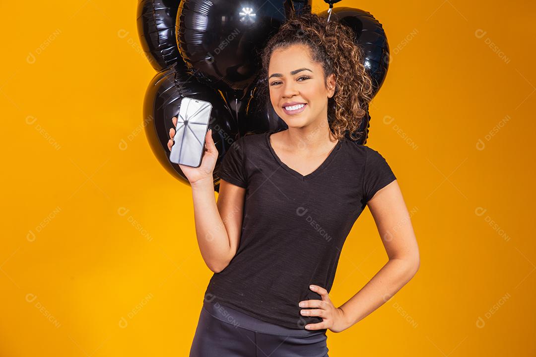 Sorrindo encantadora jovem afro segurando um celular com tela em branco vazia sobre fundo amarelo