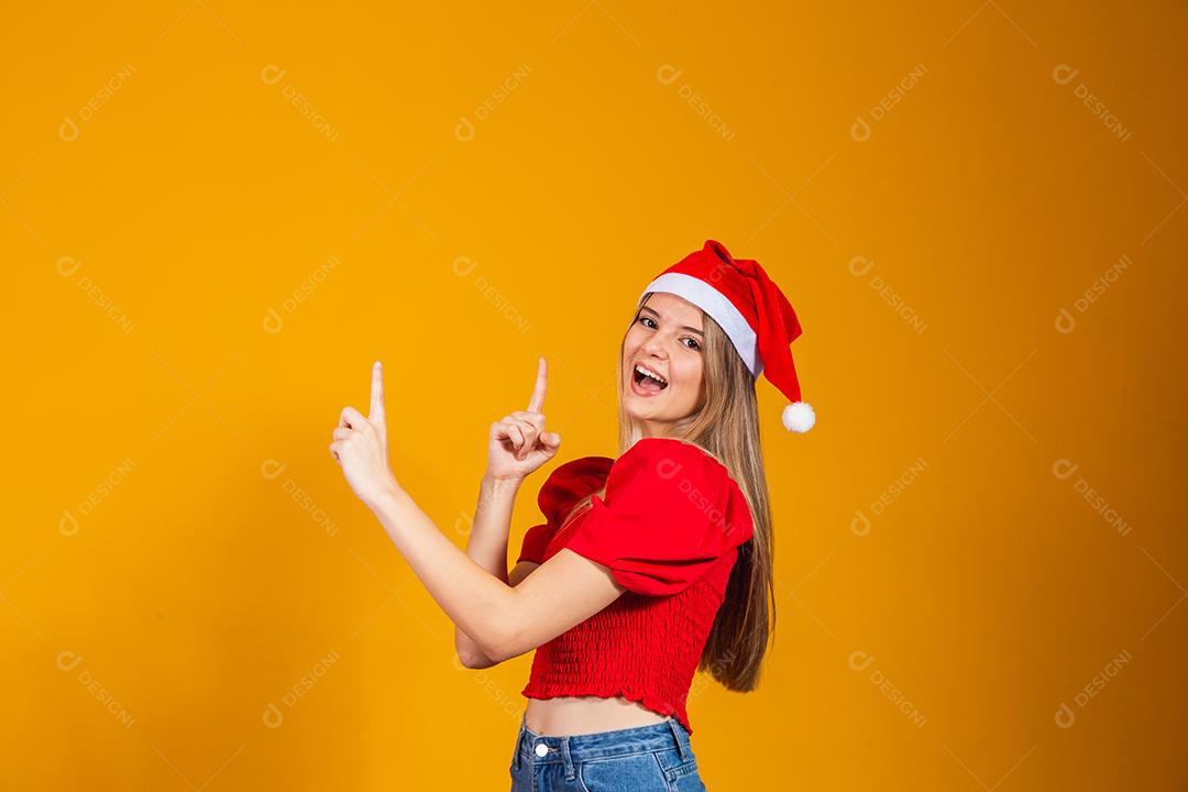 Mulher com roupa vermelha e gorro de natal apontando para cima em fundo amarelo