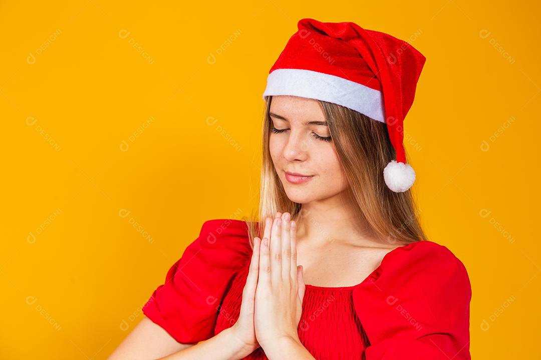 Jovem mulher bonita usando chapéu de Papai Noel Natal sobre fundo amarelo isolado, rezando com as mãos juntas