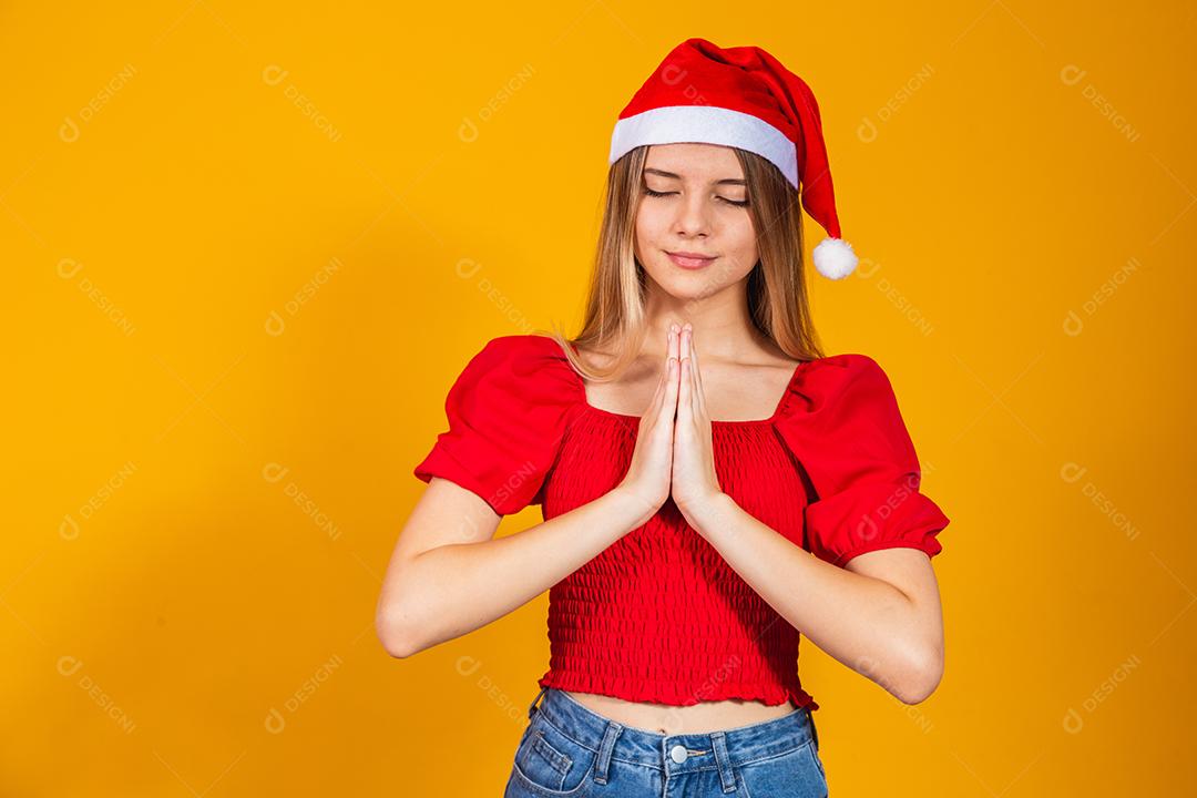 Jovem mulher bonita usando chapéu de Papai Noel Natal sobre fundo amarelo isolado, rezando com as mãos juntas