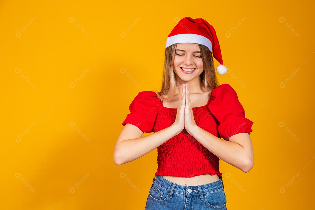 Jovem mulher bonita usando chapéu de Papai Noel Natal sobre fundo amarelo isolado, rezando com as mãos juntas