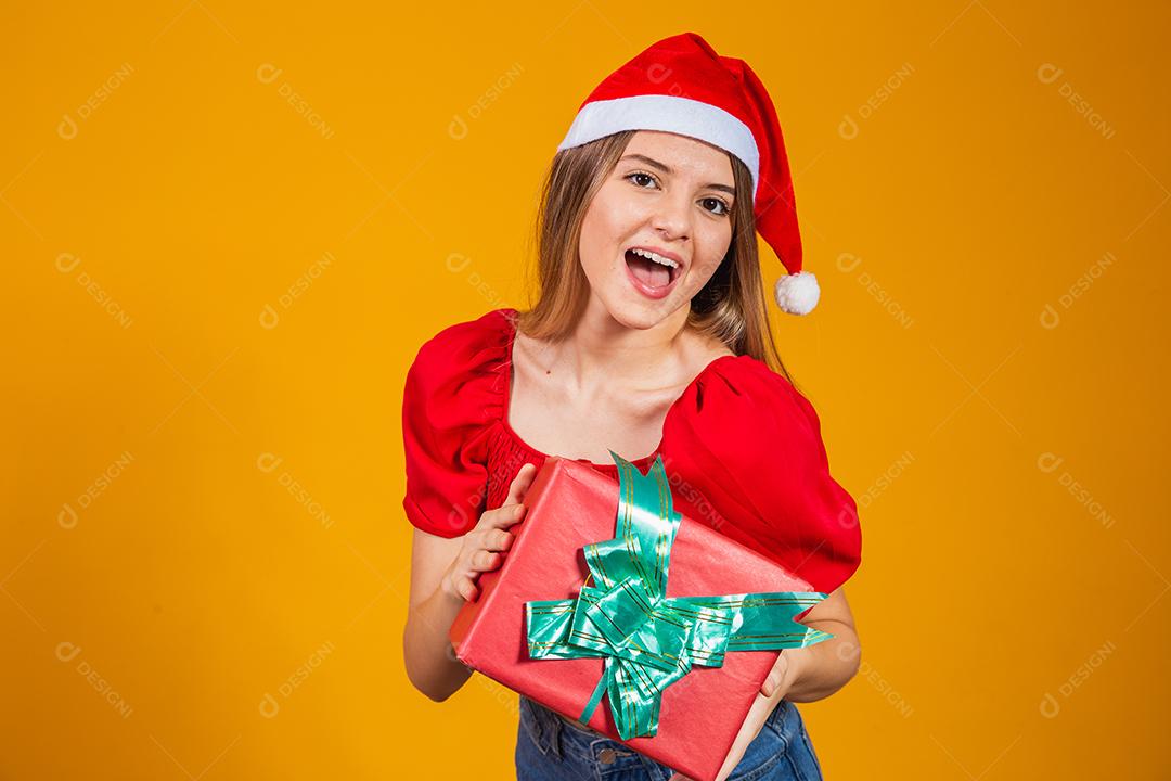 Mulher excitada sorridente com roupa de papai noel vermelha segurando presente de natal em seu ouvido isolado no fundo amarelo