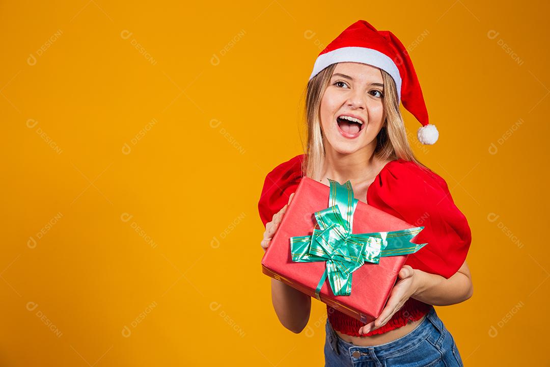 Mulher excitada sorridente com roupa de papai noel vermelha segurando presente de natal em seu ouvido isolado no fundo amarelo