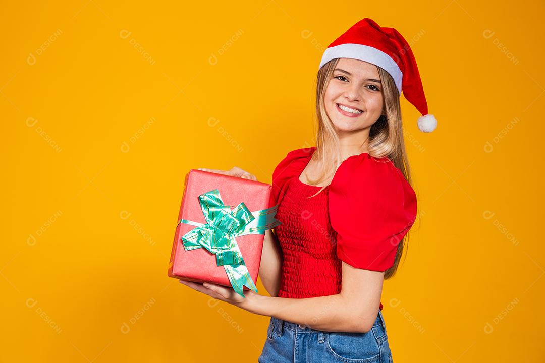 Mulher excitada sorridente com roupa de papai noel vermelha segurando presente de natal em seu ouvido isolado no fundo amarelo