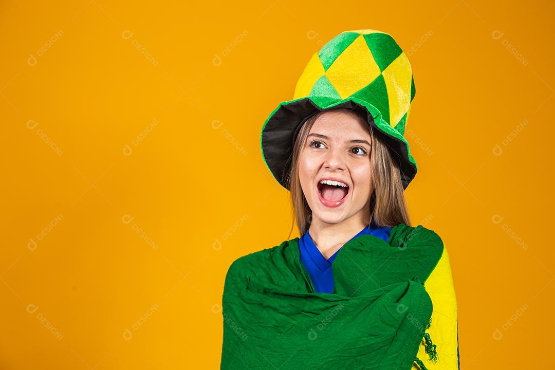 Mulher Torcedora do brasil com chapéu e bandeira e expressão de felicidade em fundo amarelo