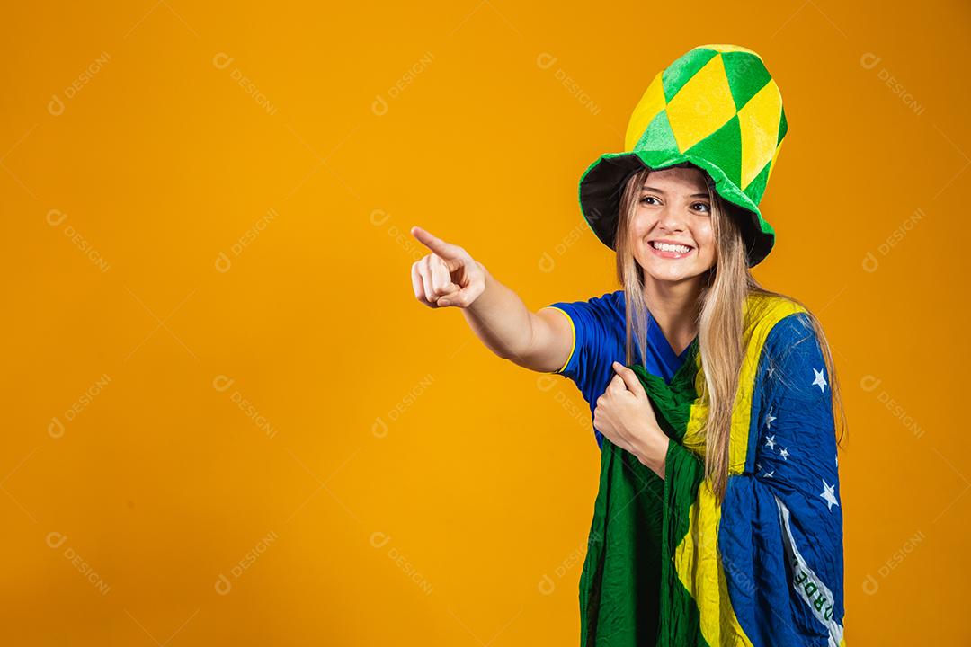Mulher apontando Torcedora do brasil com chapéu e bandeira e expressão de felicidade em fundo amarelo