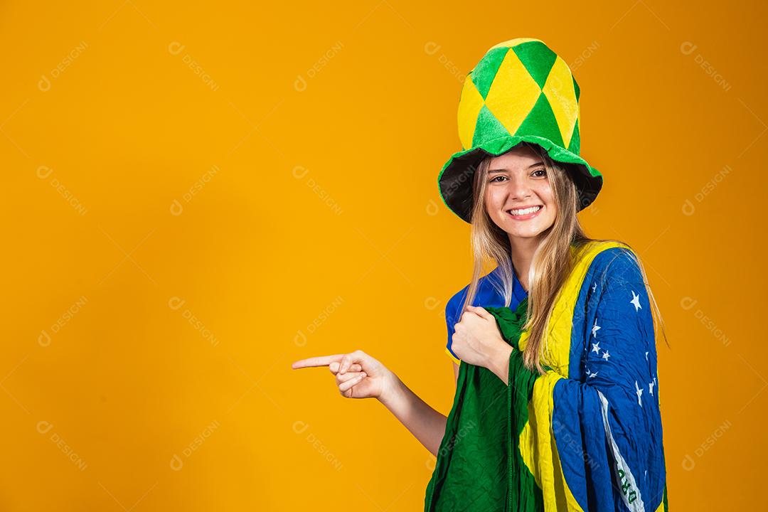 Mulher Torcedora do brasil apontando com chapéu e bandeira e expressão de felicidade em fundo amarelo