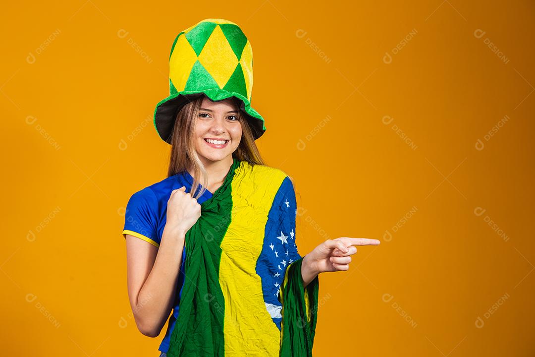 Mulher apontando Torcedora do brasil com chapéu e bandeira e expressão de felicidade em fundo amarelo