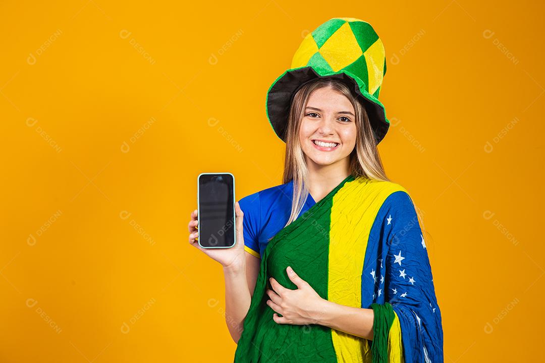 Mulher segurando celular Torcedora do brasil com chapéu e bandeira e expressão de felicidade em fundo amarelo