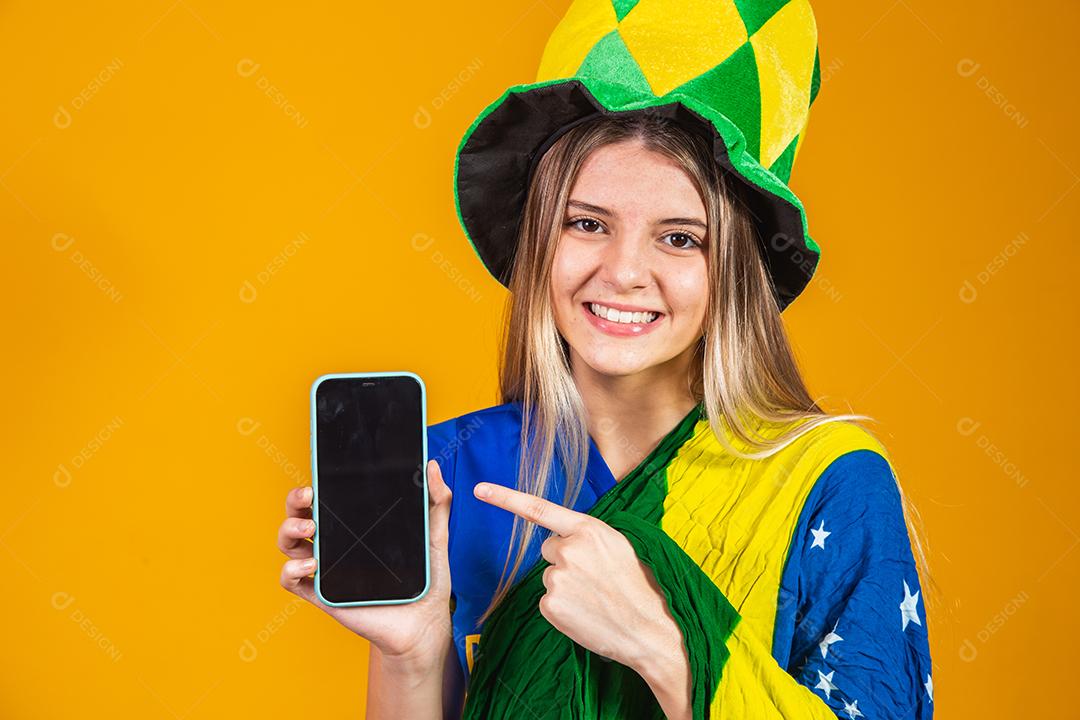 Mulher segurando celular Torcedora do brasil com chapéu e bandeira e expressão de felicidade em fundo amarelo