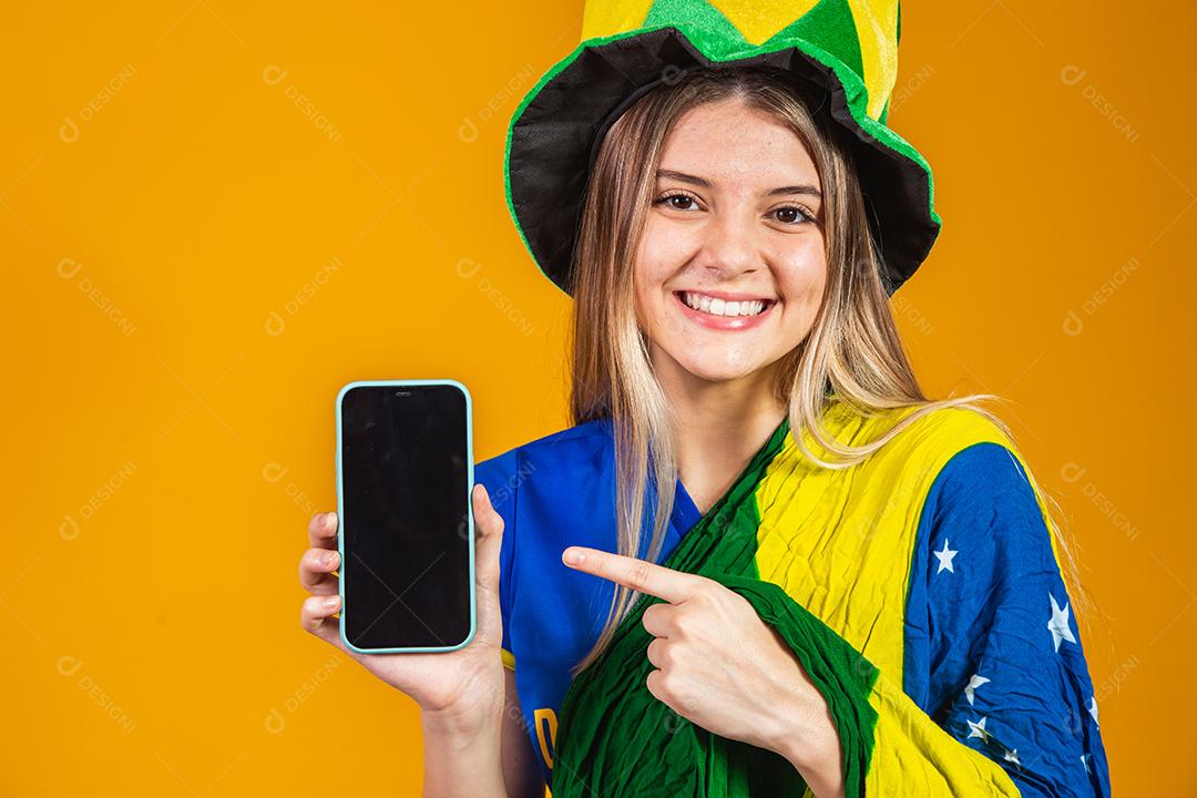 Mulher segurando celular Torcedora do brasil com chapéu e bandeira e expressão de felicidade em fundo amarelo