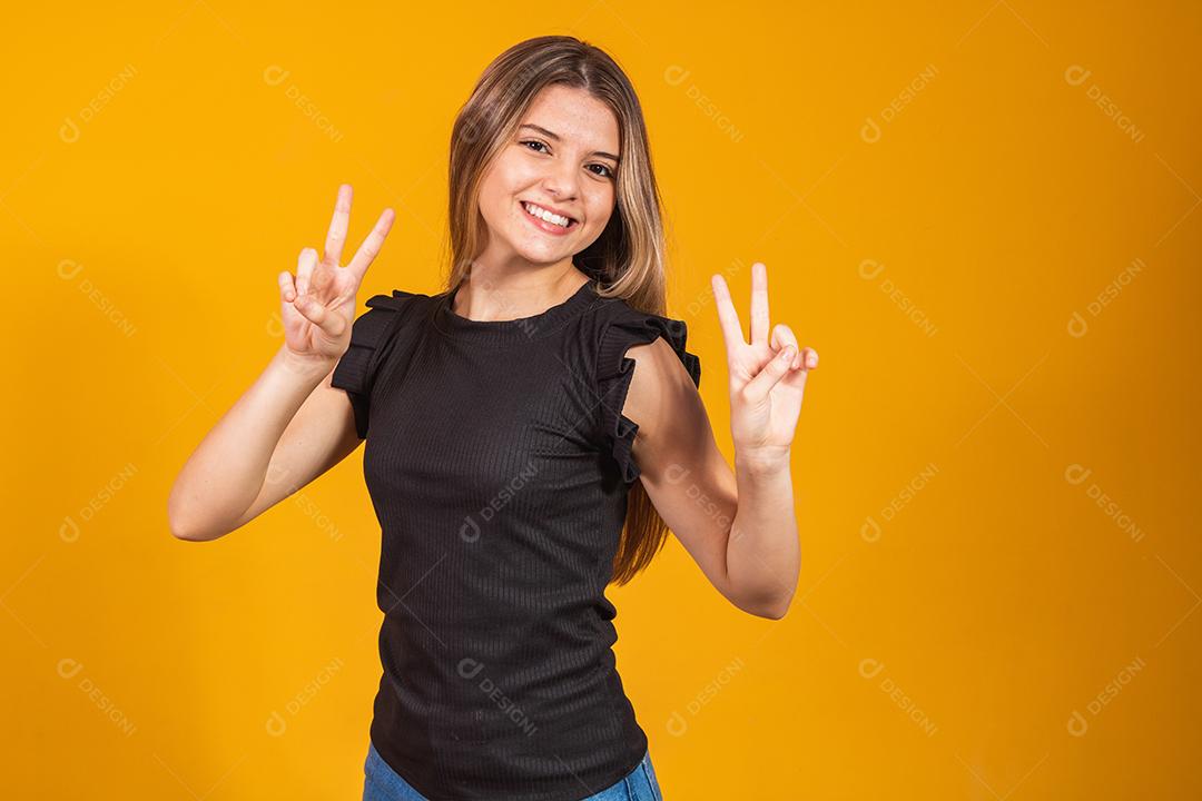 Imagem de jovem feliz em pé isolado sobre fundo amarelo, mostrando o gesto de paz. Câmera olhando.