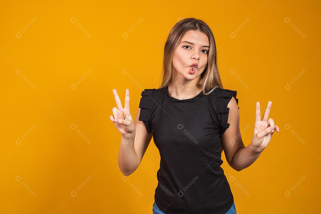 Imagem de jovem feliz em pé isolado sobre fundo amarelo, mostrando o gesto de paz. Câmera olhando.