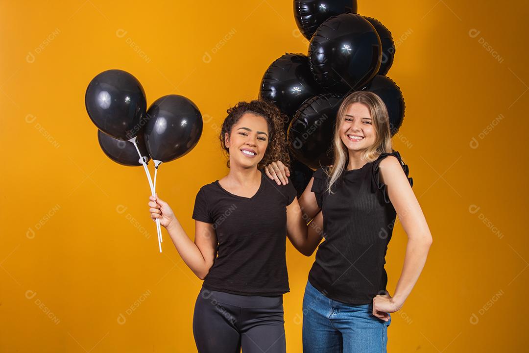 Duas namoradas em fundo amarelo com balões pretos na festa de promoção de sexta-feira negra.