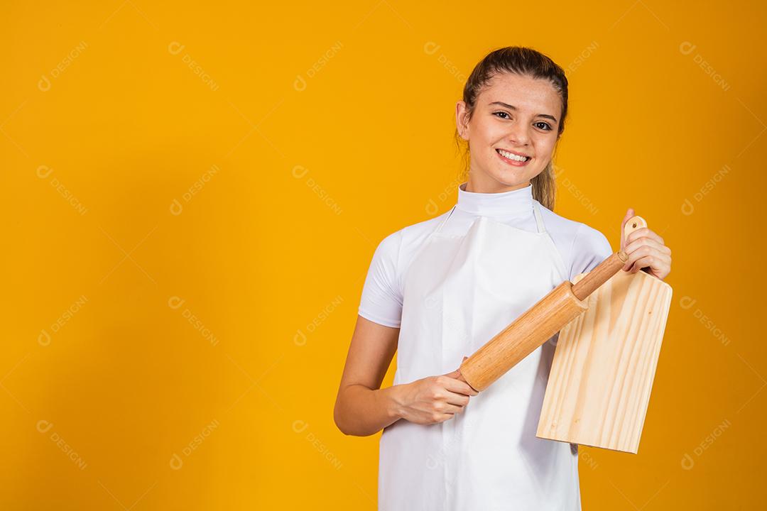 jovem mulher caucasiana com avental, tábua de carne e rolo em fundo amarelo. Cozinhar