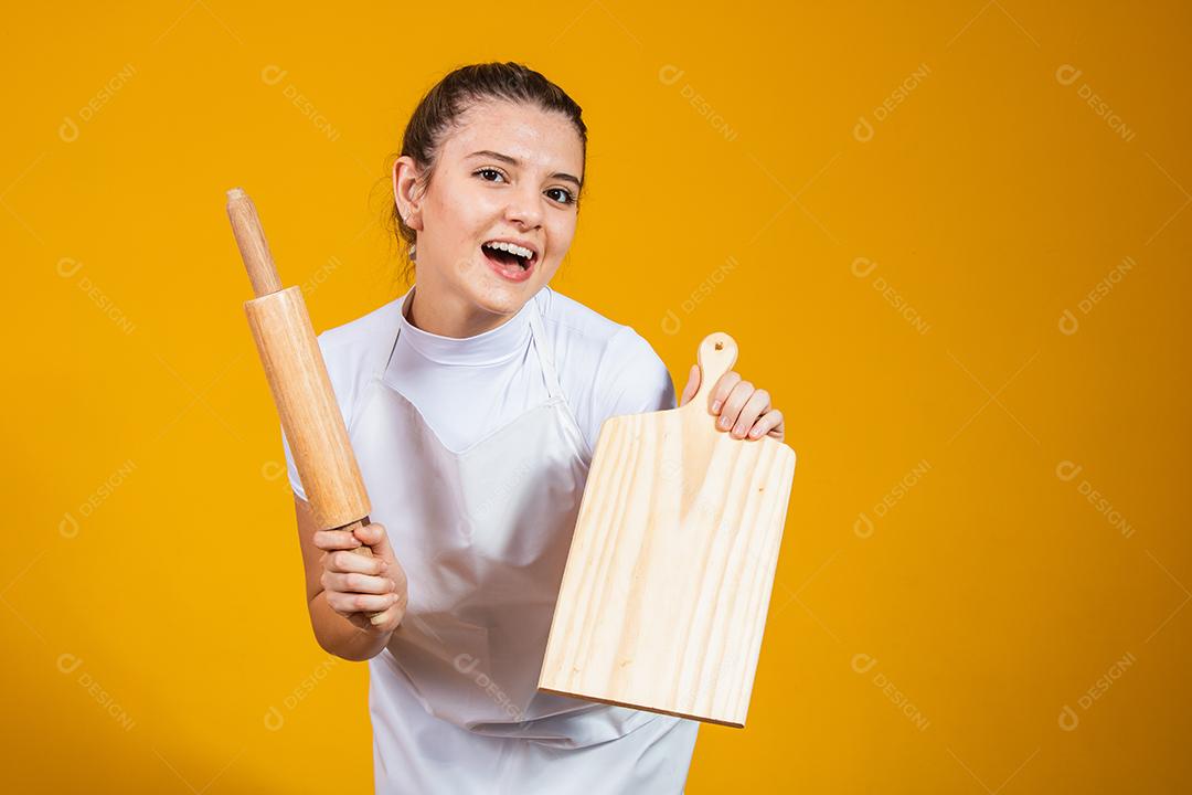 jovem mulher caucasiana com avental, tábua de carne e rolo em fundo amarelo. Cozinhar