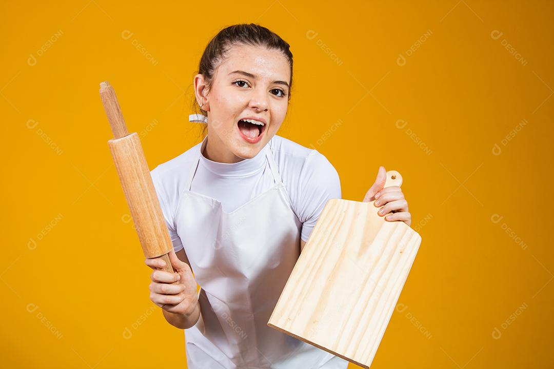 jovem mulher caucasiana com avental, tábua de carne e rolo em fundo amarelo. Cozinhar