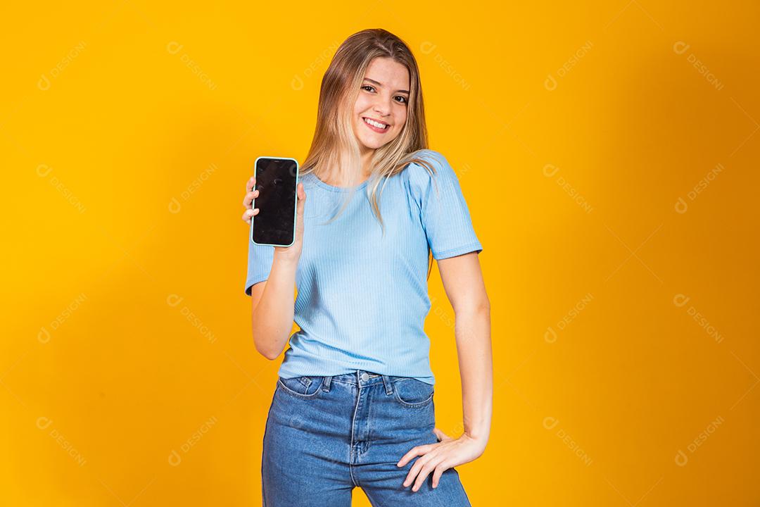 Jovem loira mostrando a tela do smartphone em branco sobre fundo amarelo.