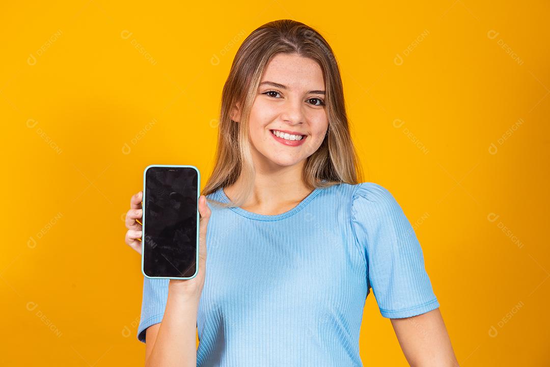 Jovem loira mostrando a tela do smartphone em branco sobre fundo amarelo.