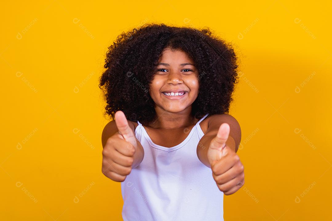 Linda garotinha de cabelos encaracolados usando as mãos okey sinal. Fundo amarelo. Garota afro ok polegar para cima