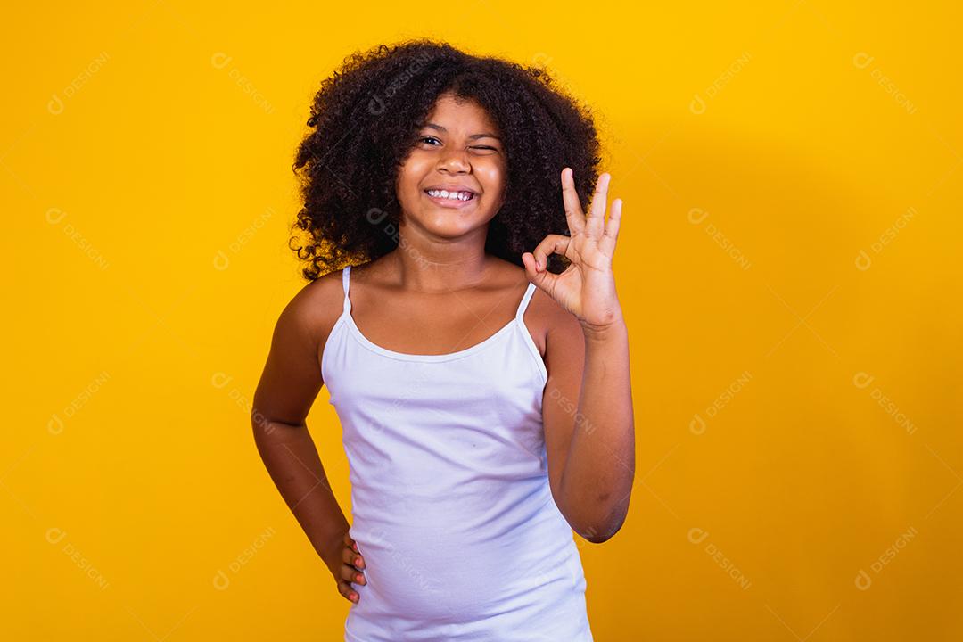 Linda garotinha de cabelos encaracolados usando as mãos okey sinal. Fundo amarelo. mulher afro ok