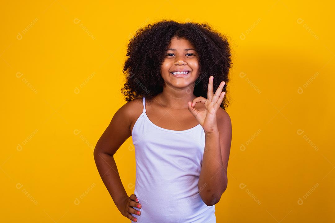 Linda garotinha de cabelos encaracolados usando as mãos okey sinal. Fundo amarelo. Garota afro ok polegar para cima