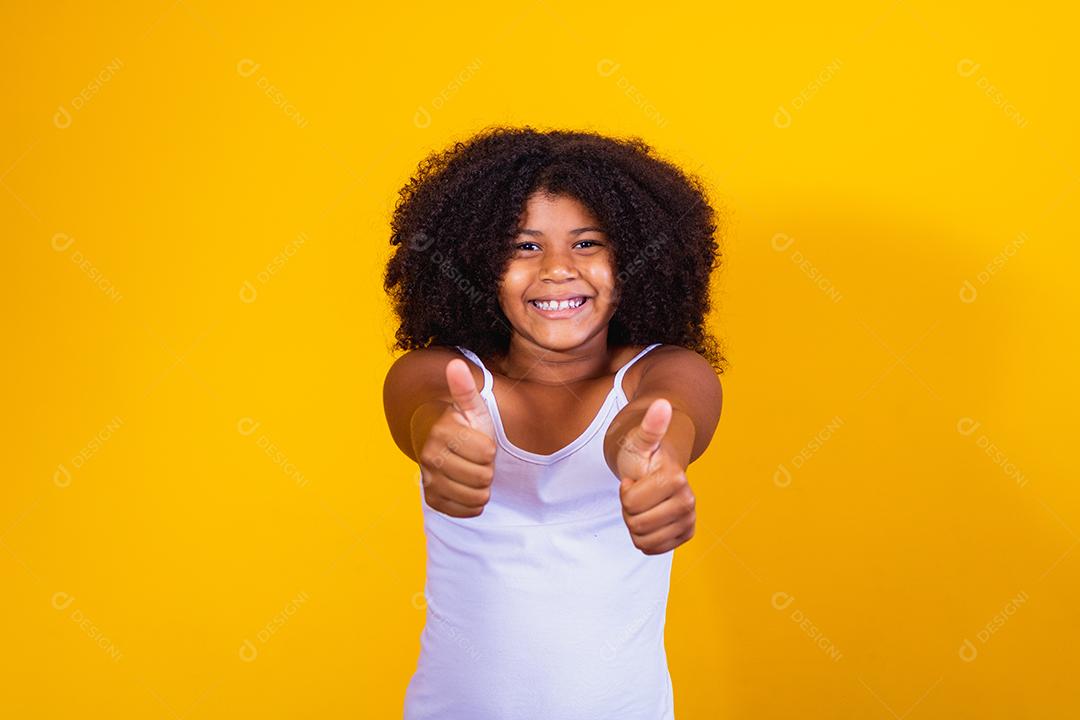 Linda garotinha de cabelos encaracolados usando as mãos okey sinal. Fundo amarelo. Garota afro ok polegar para cima