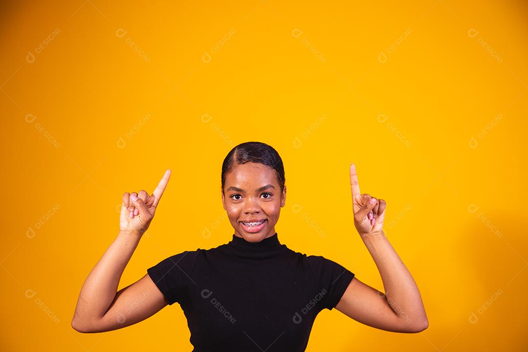 Mulher afro sorrindo em fundo amarelo, apontando para cima