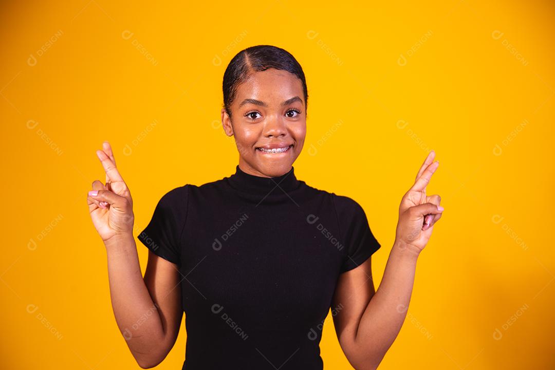 Jovem e bela mulher afro, elegante, gesticulando com o dedo cruzado, sorrindo com esperança e olhos fechados