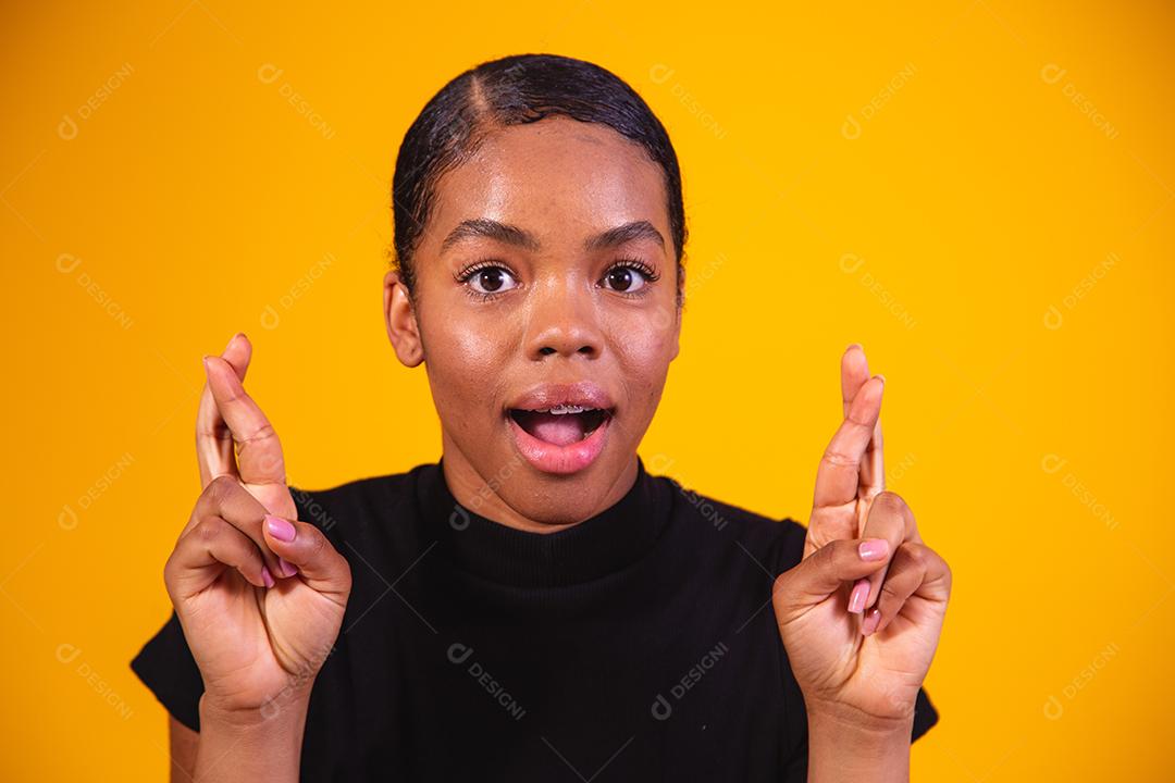 Jovem e bela mulher afro, elegante, gesticulando com o dedo cruzado, sorrindo com esperança e olhos fechados