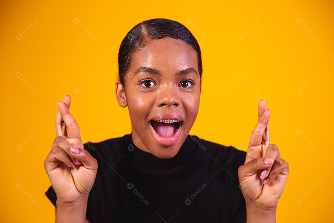 Jovem e bela mulher afro, elegante, gesticulando com o dedo cruzado, sorrindo com esperança e olhos fechados