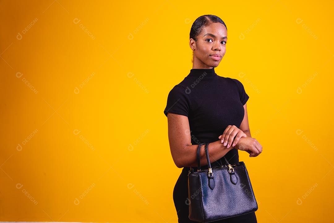 Mulher de negócios em fundo amarelo, vestindo roupas pretas e bolsa de mão.