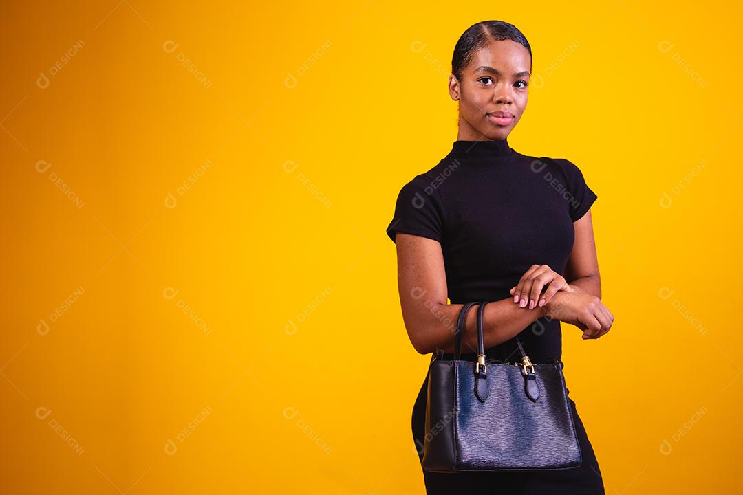 Mulher de negócios em fundo amarelo, vestindo roupas pretas e bolsa de mão.