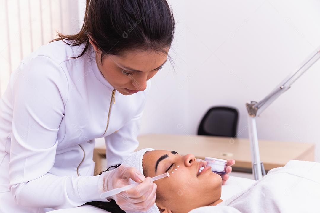 mulher bonita com máscara facial no salão de beleza. Esteticista aplicando creme no rosto do paciente