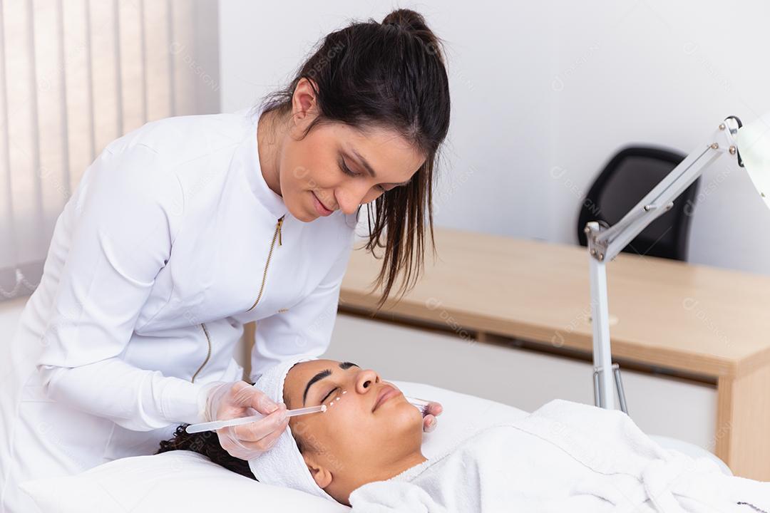 mulher bonita com máscara facial no salão de beleza. Esteticista aplicando creme no rosto do paciente