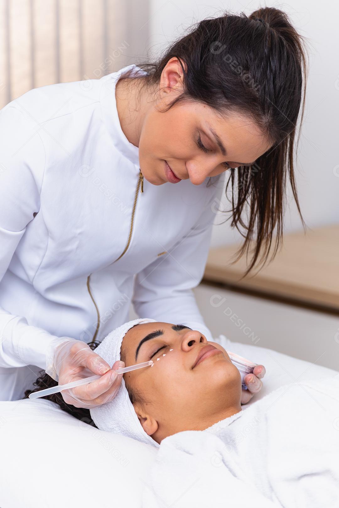 mulher bonita com máscara facial no salão de beleza. Esteticista aplicando creme no rosto do paciente