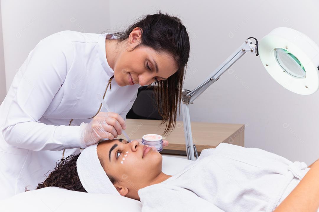 mulher bonita com máscara facial no salão de beleza. Esteticista aplicando creme no rosto do paciente